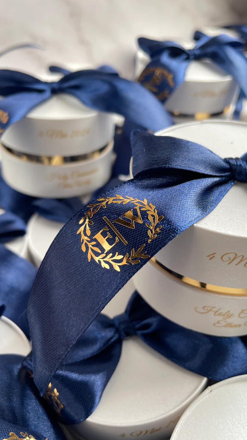 Personalized round ivory wedding favor boxes with navy blue satin bows and gold monogram details for Nikkah and Pakistani wedding celebrations.