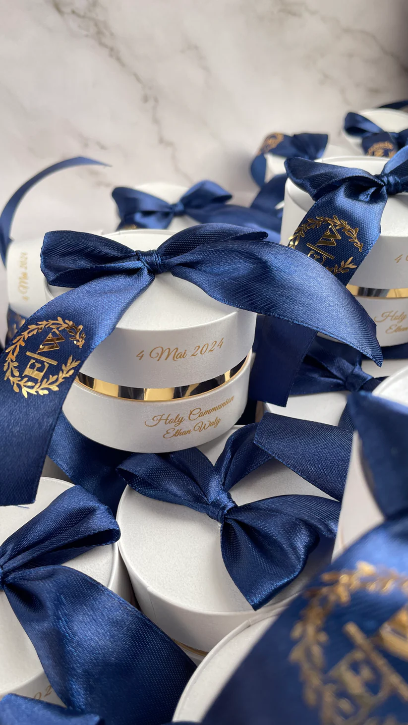 Personalized round ivory wedding favor boxes with navy blue satin bows and gold monogram details for Nikkah and Pakistani wedding celebrations.