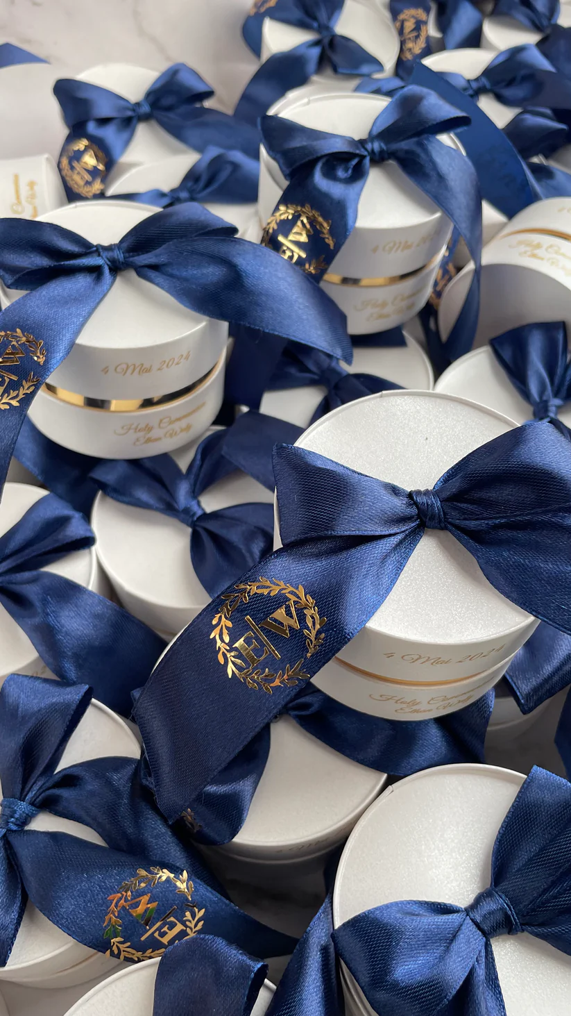 Personalized round ivory wedding favor boxes with navy blue satin bows and gold monogram details for Nikkah and Pakistani wedding celebrations.