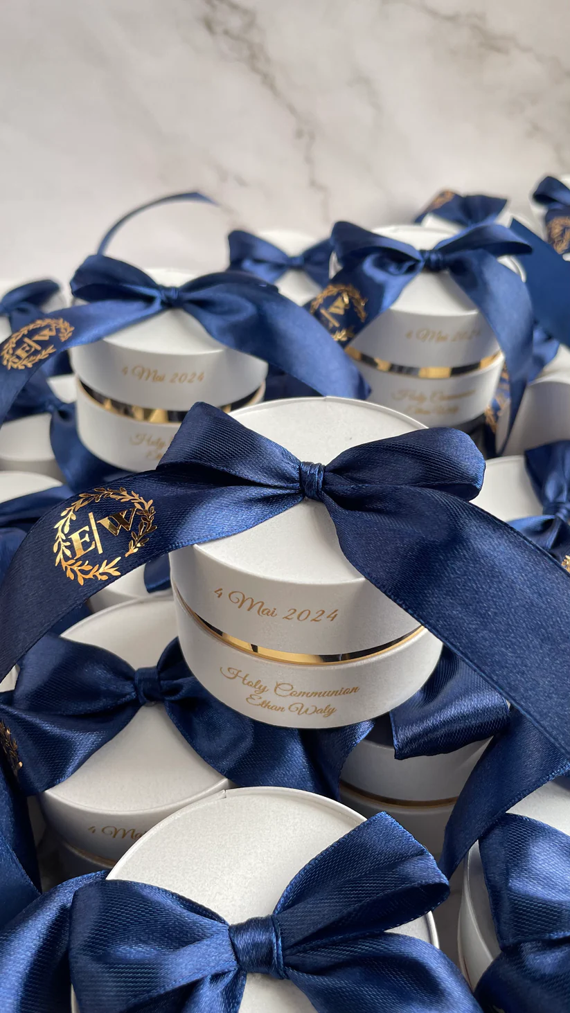Personalized round ivory wedding favor boxes with navy blue satin bows and gold monogram details for Nikkah and Pakistani wedding celebrations.