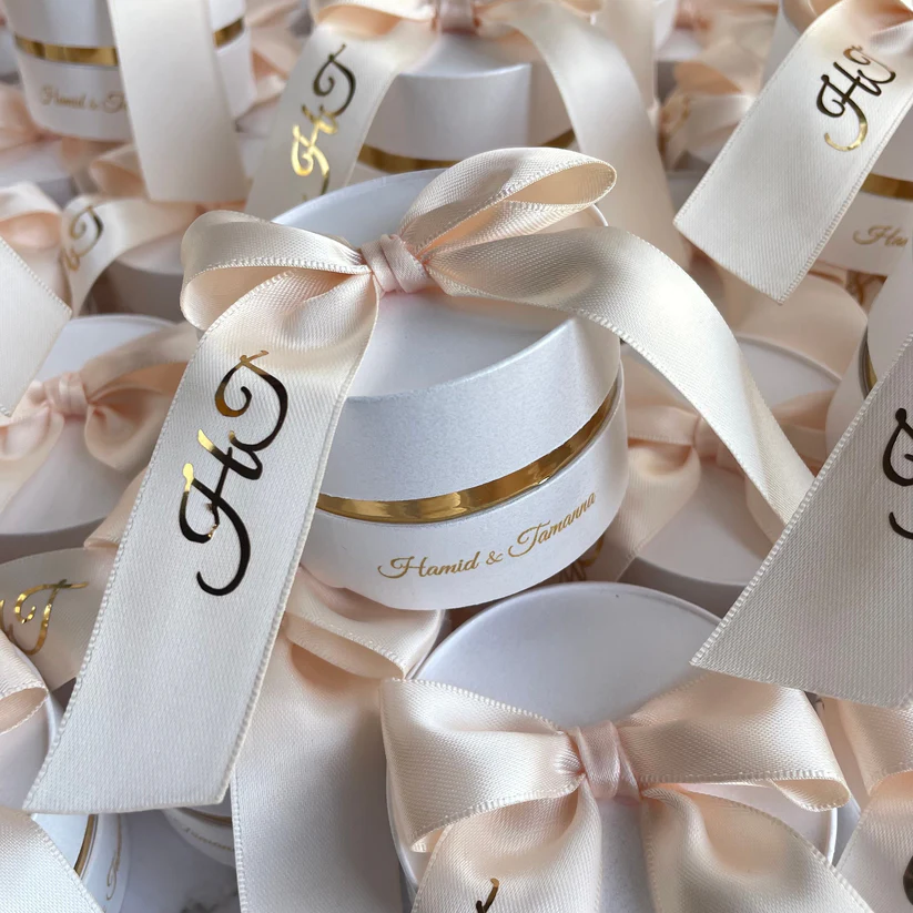 Personalized round ivory Nikkah favor boxes with custom satin initial ribbons and elegant gold accents for Pakistani and Muslim weddings.