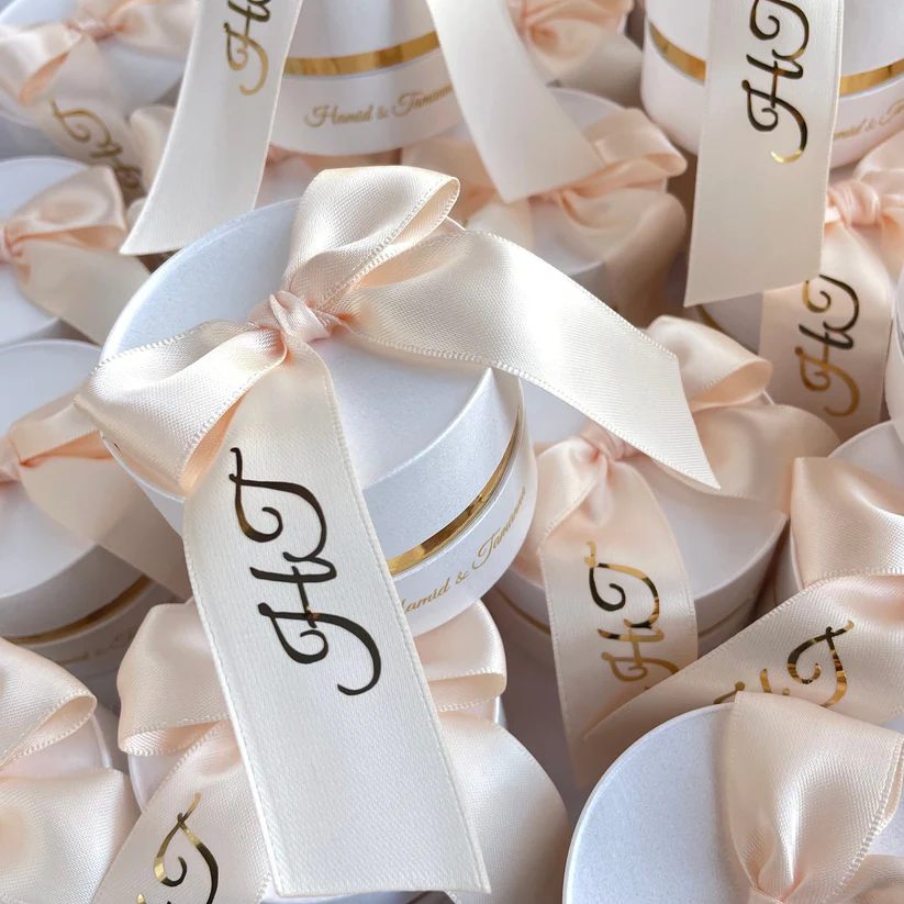 Personalized round ivory Nikkah favor boxes with custom satin initial ribbons and elegant gold accents for Pakistani and Muslim weddings.