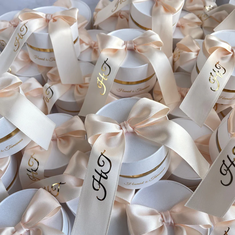 Personalized round ivory Nikkah favor boxes with custom satin initial ribbons and elegant gold accents for Pakistani and Muslim weddings.