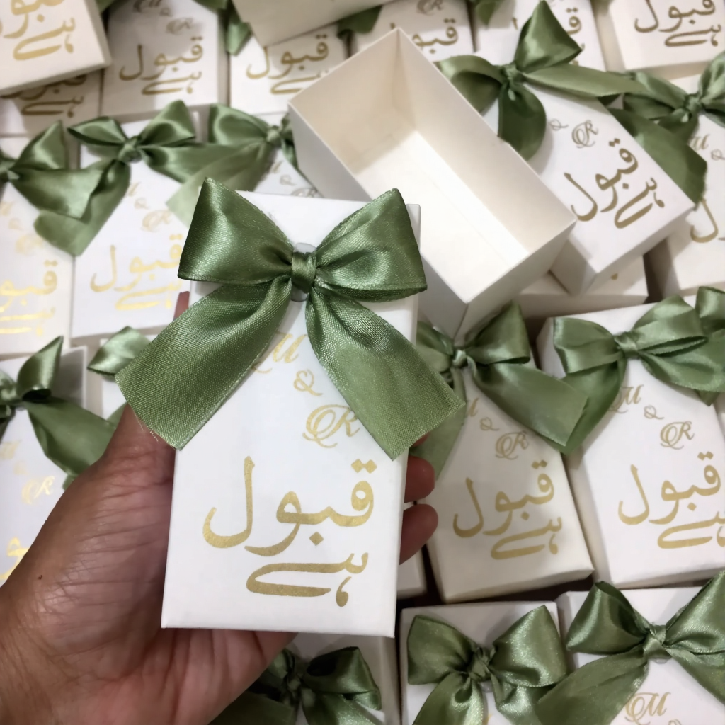 Ivory Qabool Hai Nikkah favor boxes with gold Urdu calligraphy and sage green satin bows for Pakistani and Muslim weddings.