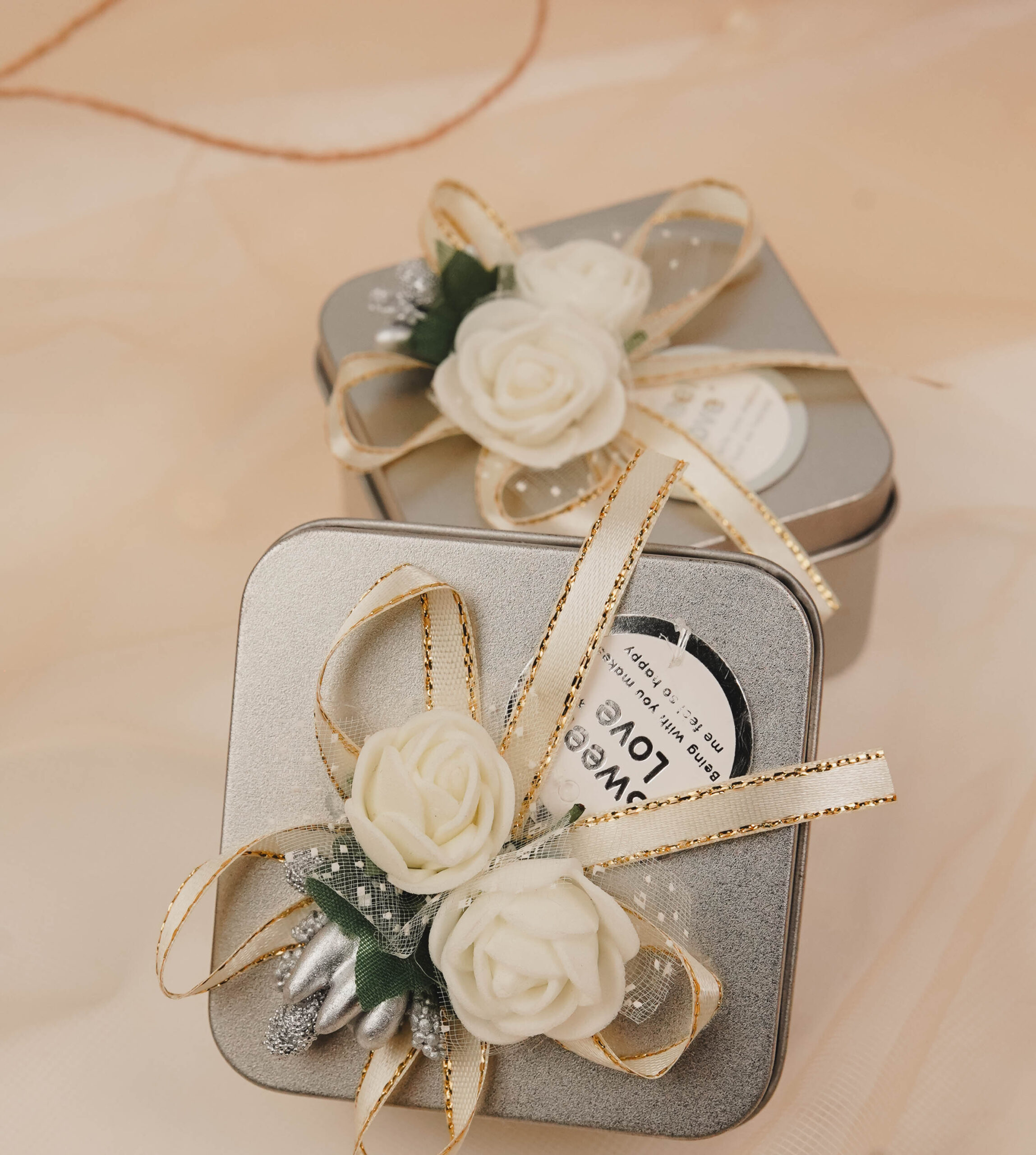 Silver floral tin favor boxes decorated with ivory flowers and gold-trimmed ribbons for Nikkah, Pakistani weddings and engagement celebrations.
