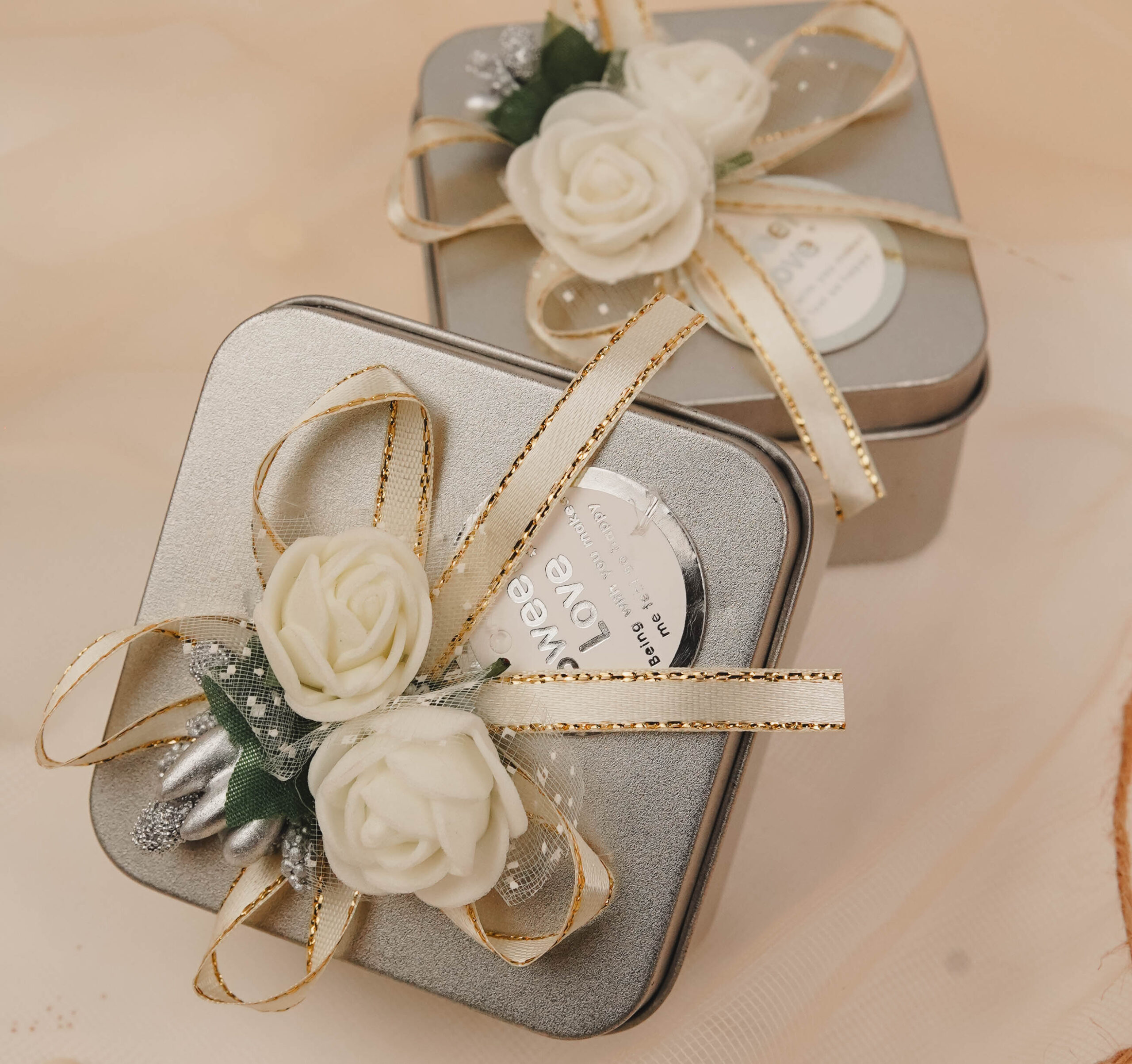 Silver floral tin favor boxes decorated with ivory flowers and gold-trimmed ribbons for Nikkah, Pakistani weddings and engagement celebrations.