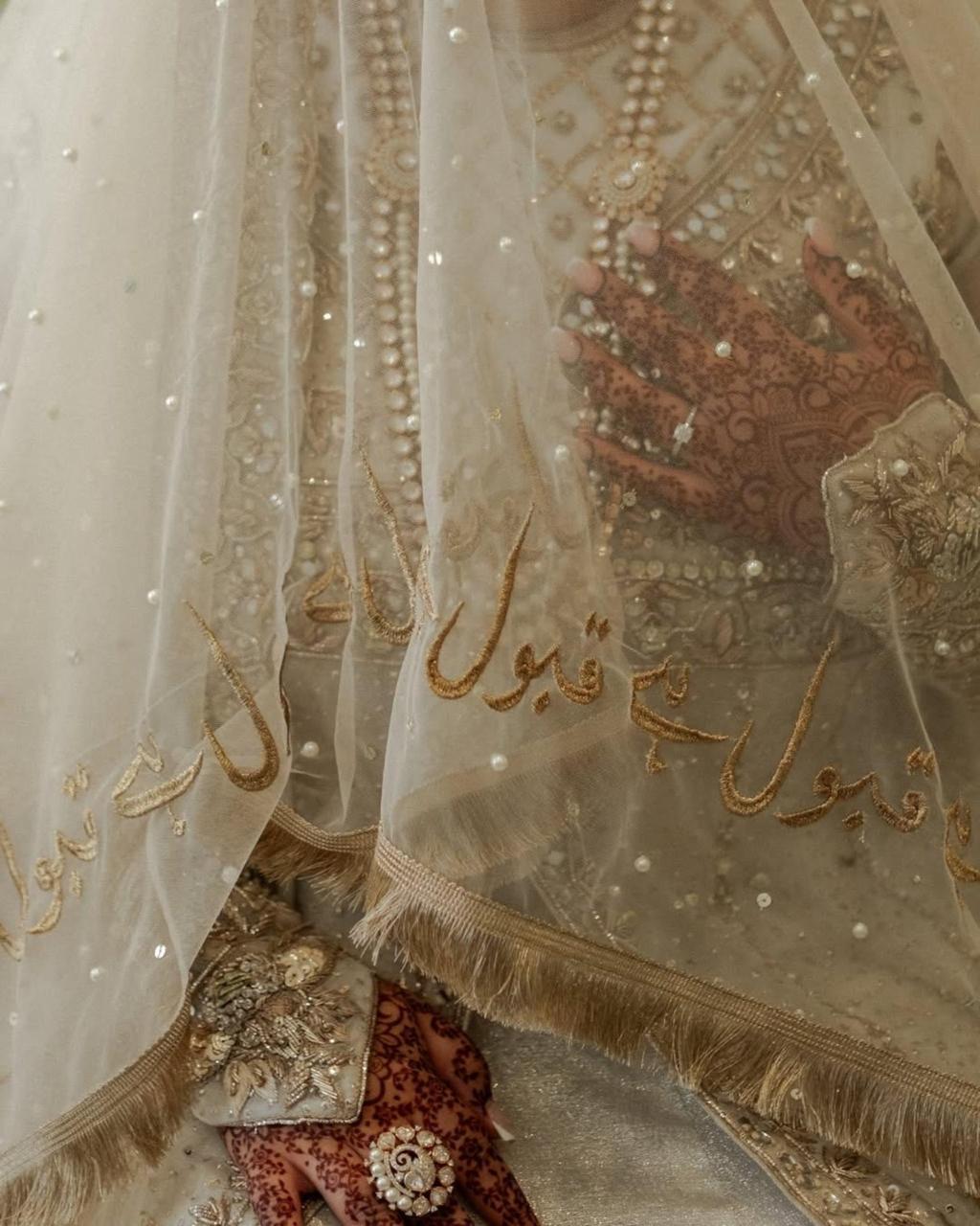 Ivory Pakistani bridal Nikkah dupatta featuring delicate sheer net fabric, gold embroidered borders, shimmering sequins, and Urdu calligraphy, elegantly draped over a Muslim bride during her wedding ceremony.