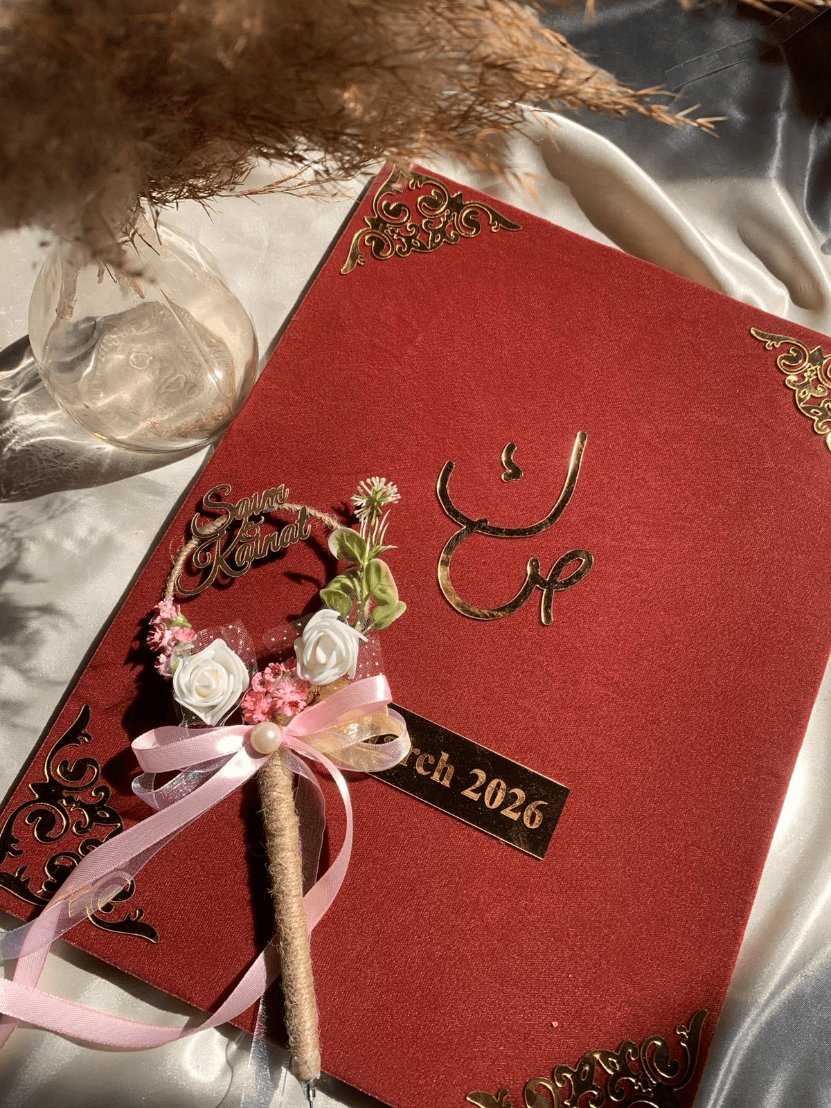 Personalized maroon velvet Nikkah Nama folder featuring metallic gold Urdu calligraphy, intricate ornamental gold corners, custom wedding date plaque, and decorative floral signing pen with pink satin ribbons for Pakistani and Muslim weddings in the USA.