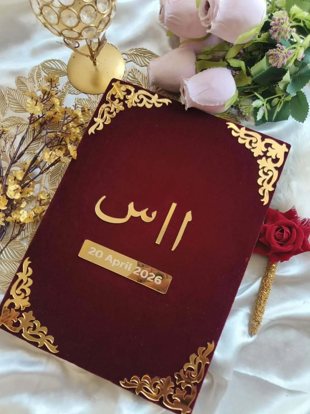 Personalized maroon velvet Nikkah Nama folder featuring metallic gold Urdu initials, a custom wedding date plaque, and ornate gold floral corner embellishments for Pakistani and Muslim wedding ceremonies.